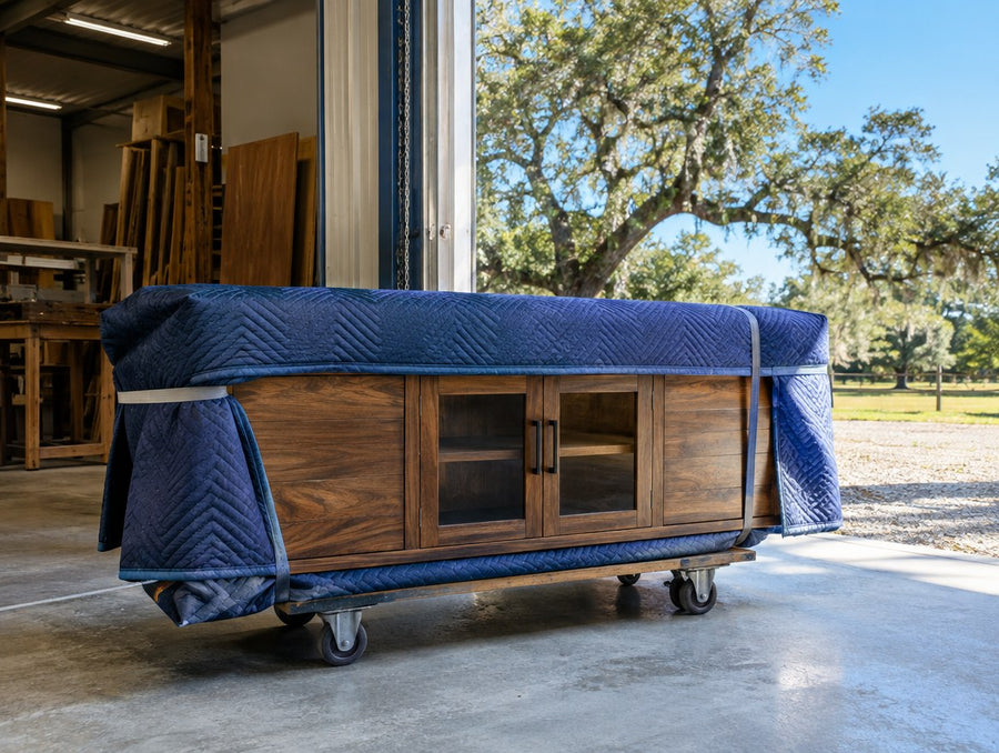 A finished solid wood TV console wrapped in moving blankets at the roll-up door of a cabinet shop